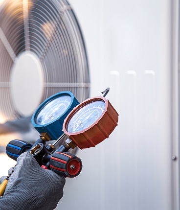 Technicien portant un gant gris vérifiant la pression d'un climatiseur extérieur avec un manomètre bleu et rouge.
