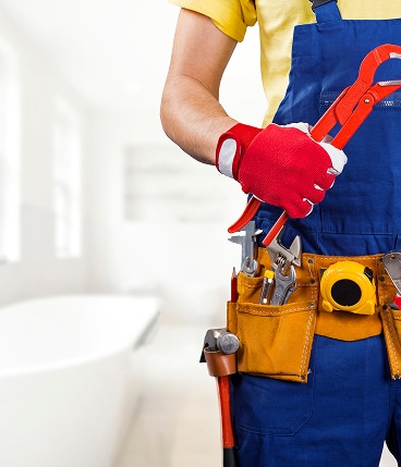 Plombier en salopette bleue et gants rouges tenant une clé serre-tube, avec ceinture porte-outils, devant une salle de bain floue.