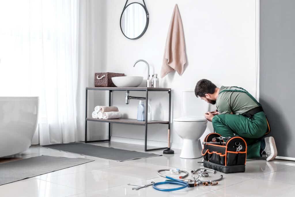 Plombier en salopette verte réparant une toilette blanche dans une salle de bain moderne avec lavabo sur meuble métallique.