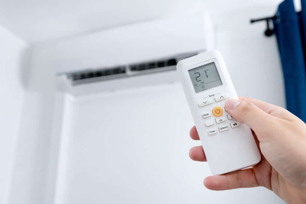 Main tenant une télécommande blanche réglant la climatisation sur 21°C en mode COOL, avec l'unité en arrière-plan.